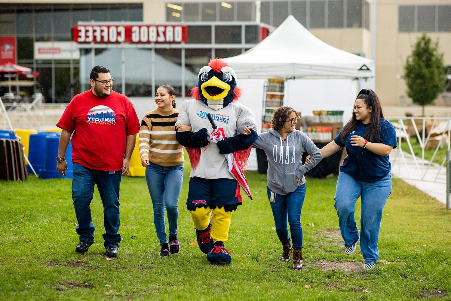 Four alumns walking with Rowdy the Roadrunner at the Homecoming Tailgate on the grass outside of the JSSB building